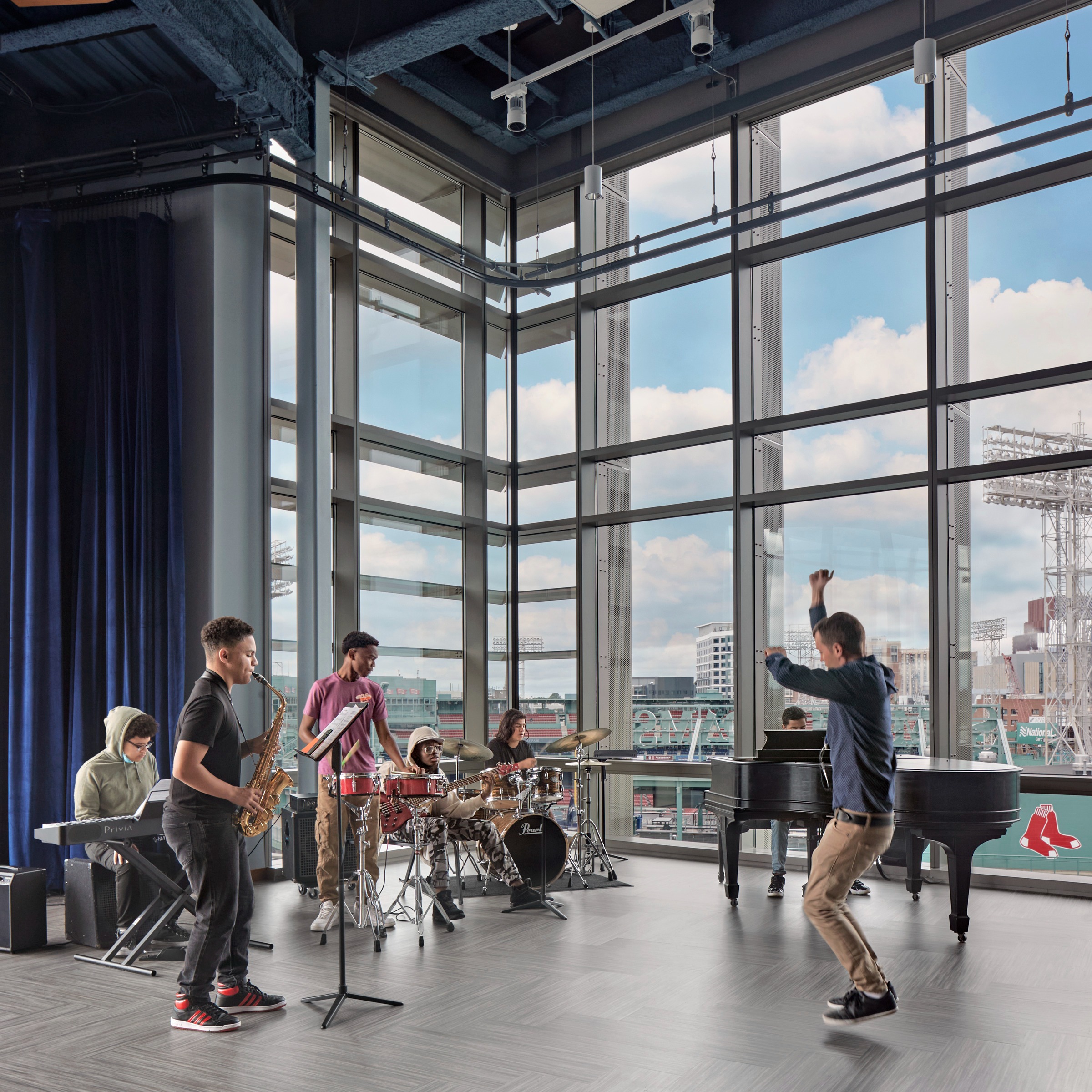 An animated band instructor jumps with enthusiasm while his students play with a background of floor to ceiling windows and blue velvet curtains. The sky beyond is blue with white puffy clouds. This building overlooks Fenway Park.