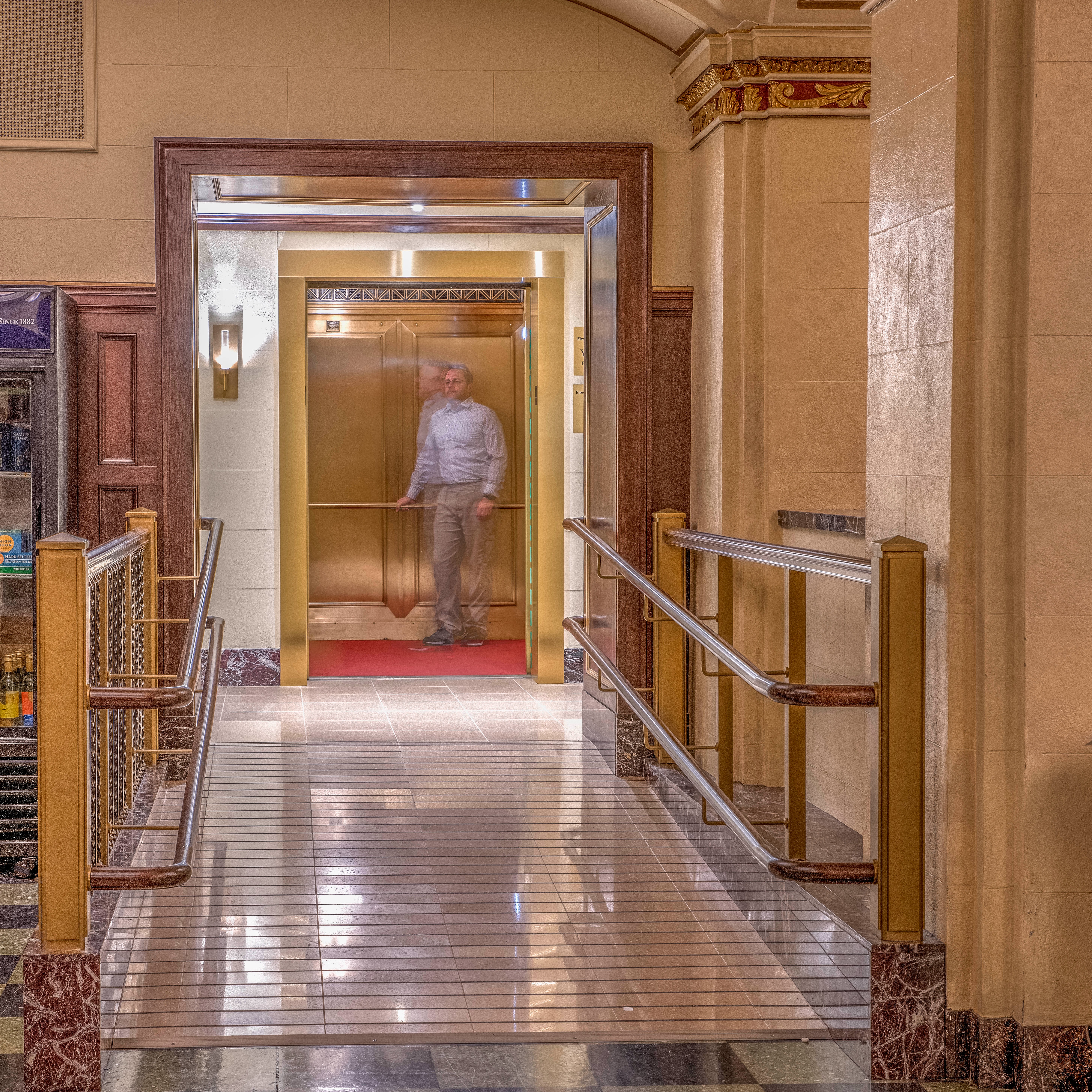 Within a historic interior, a ramp with railings leads up to an open elevator door, revealing a man standing on a red carpet surrounded by the brass walls of the historic elevator cab at the Wang Theatre.