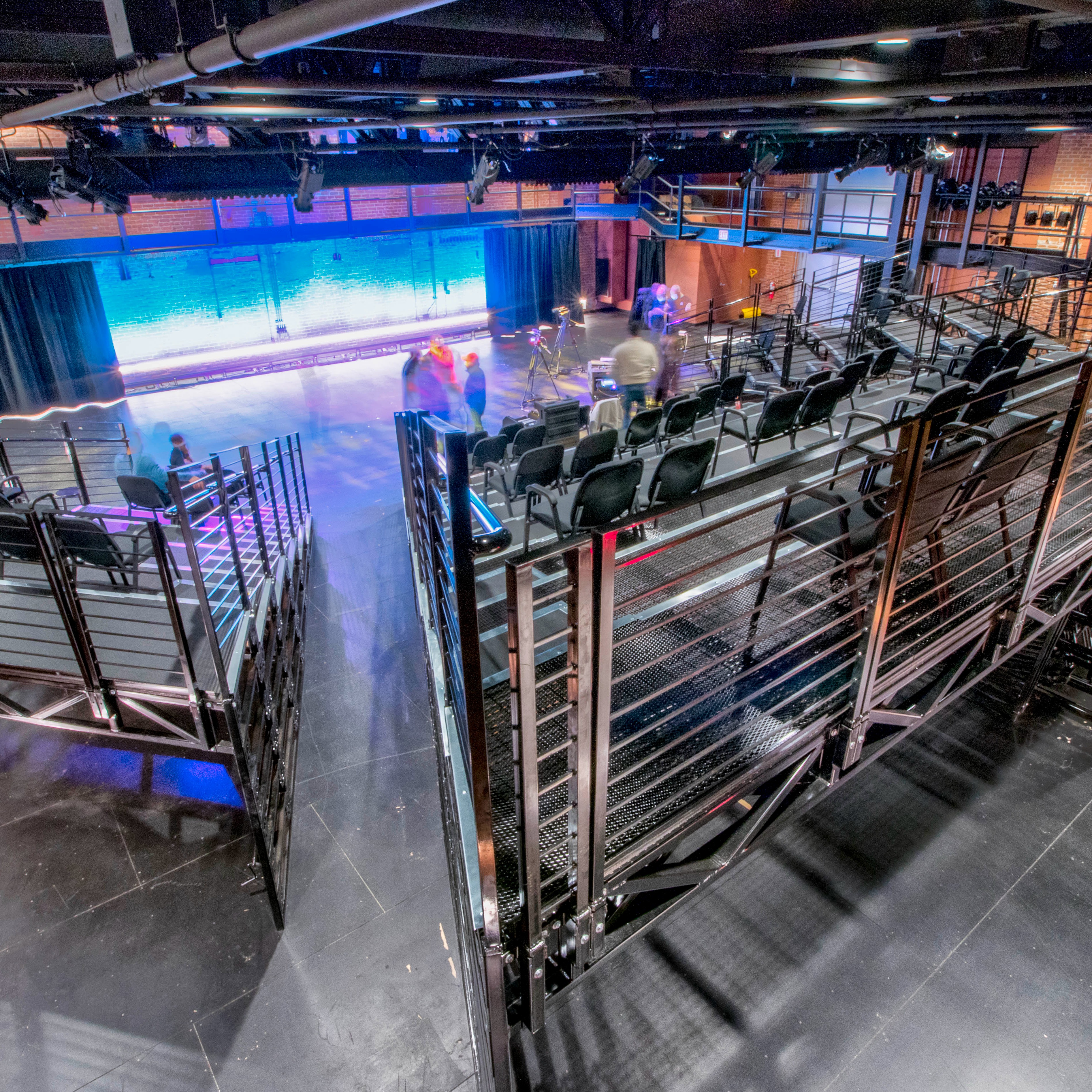 A view of a flexible black-box style space with movable seating platforms aimed towards a stage area with brick wall backdrop illuminated by blue LEDs. Some people gather in the stage area.