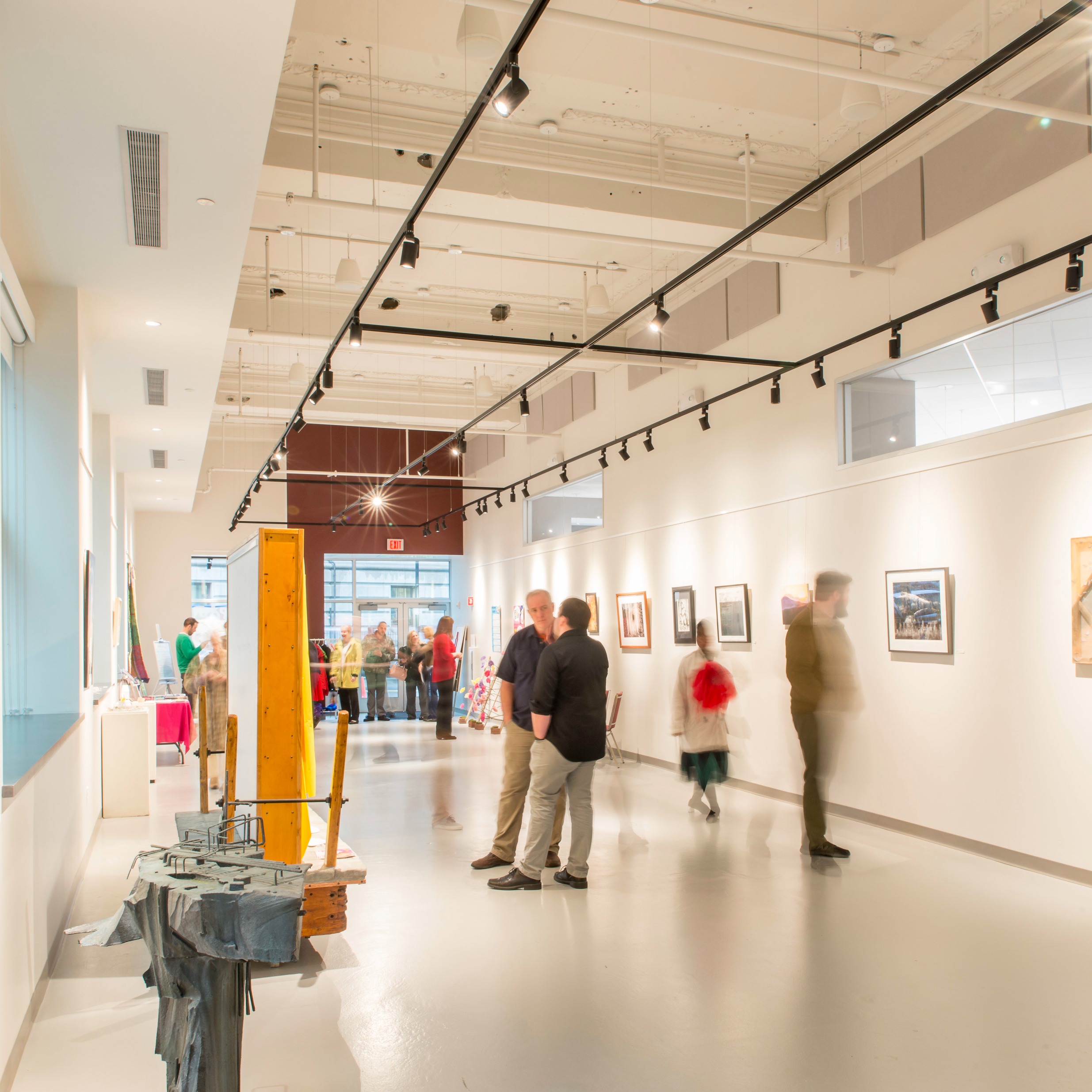 People look at art and chat in a bright white gallery space with black track lighting above. Artwork is displayed on the far wall and sculptural pieces are seen in the foreground.