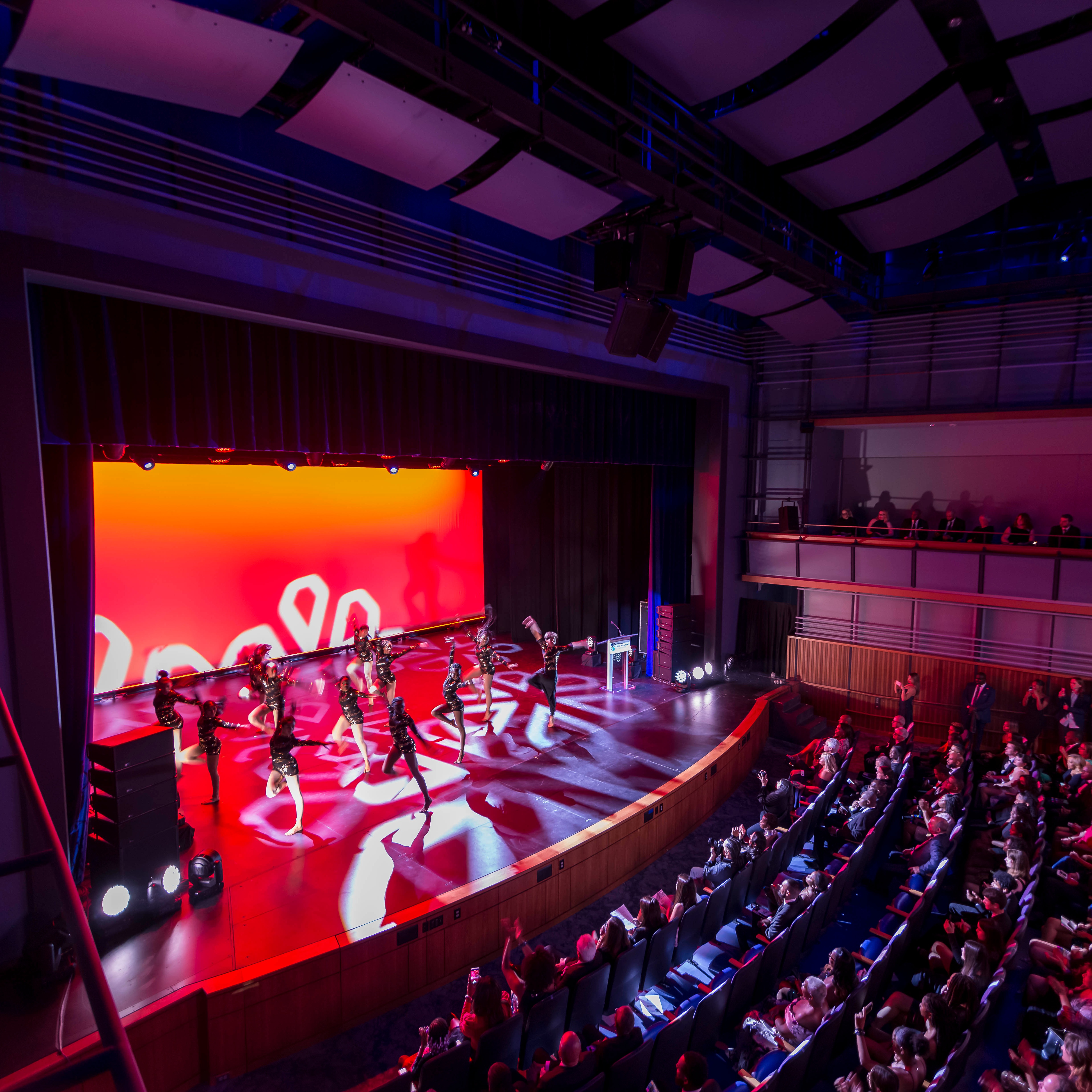 A dark theater with a bright orange-red backdrop on stage throws light around the room while a dance group performs a synchronized dance and the audience looks on.