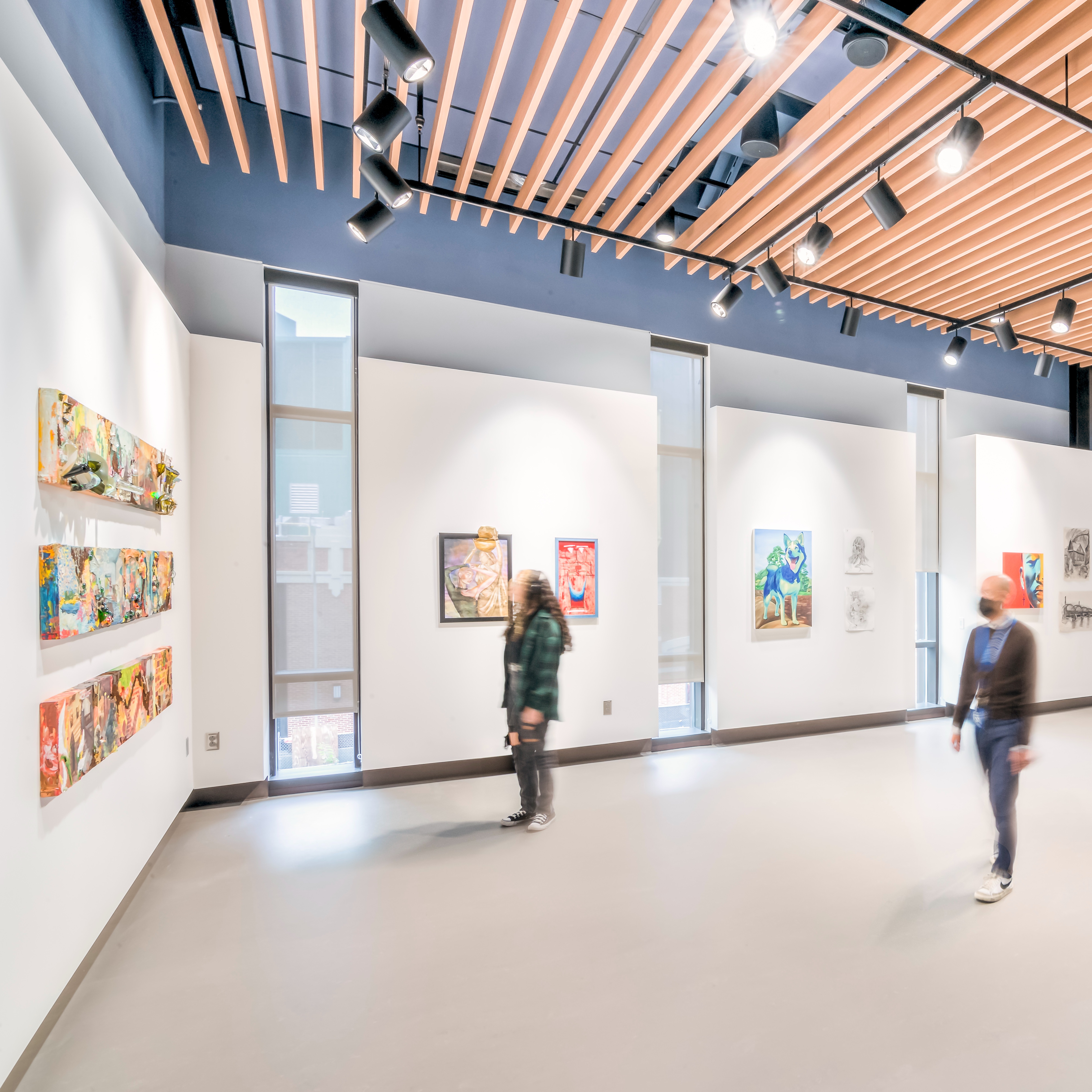 People gaze at artwork in a brightly lit gallery space, a wood slat ceiling with black track lighting overhead. Colorful fashion pieces on mannequins can be seen to the right.