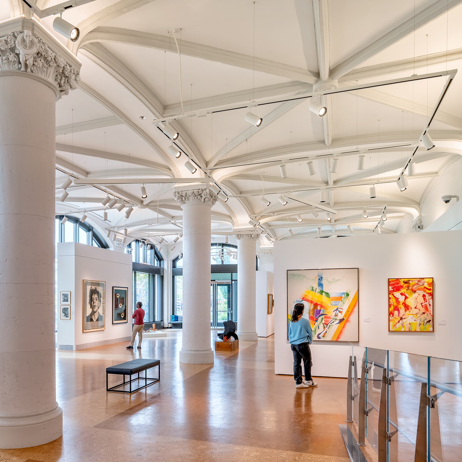 A daylit art gallery space with historic columns and vaulted ceilings. Two people admire artwork on the walls.