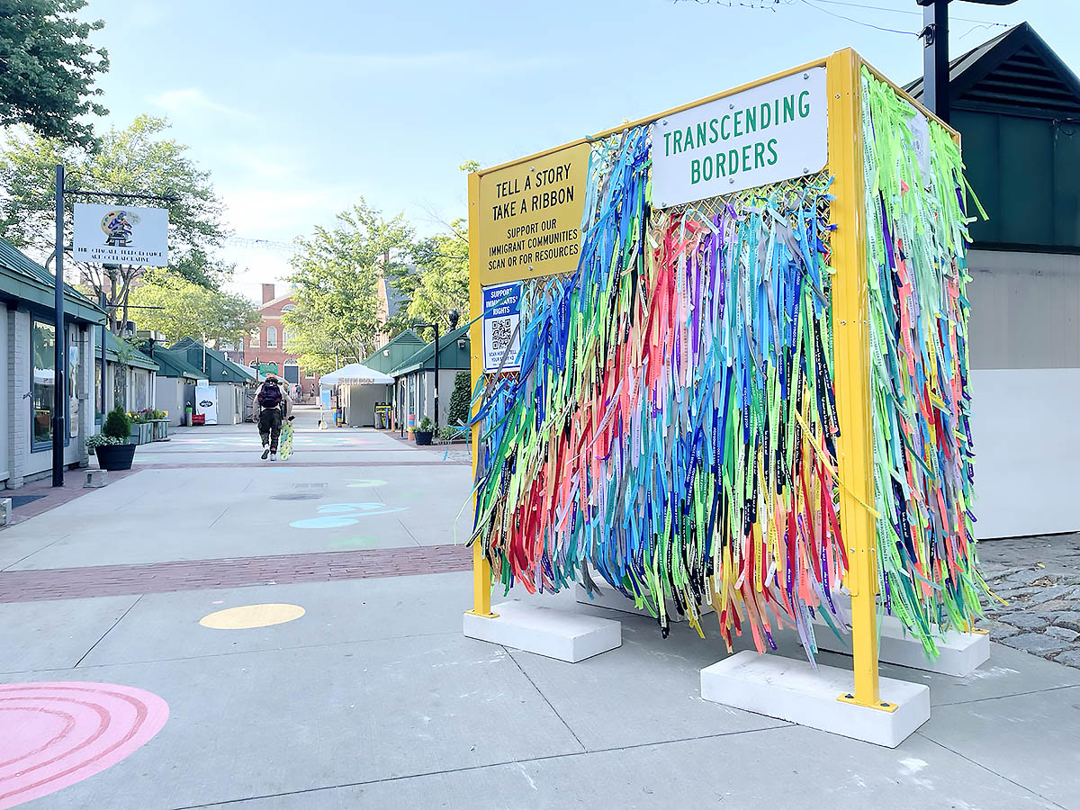 Transcending Borders Immigrant Experiences and Dreams a yellow modular fence structure covered by over 2,000 brightly colored ribbons with the phrase you are welcome here printed in 12 languages commonly spoken by immigrants in Boston 