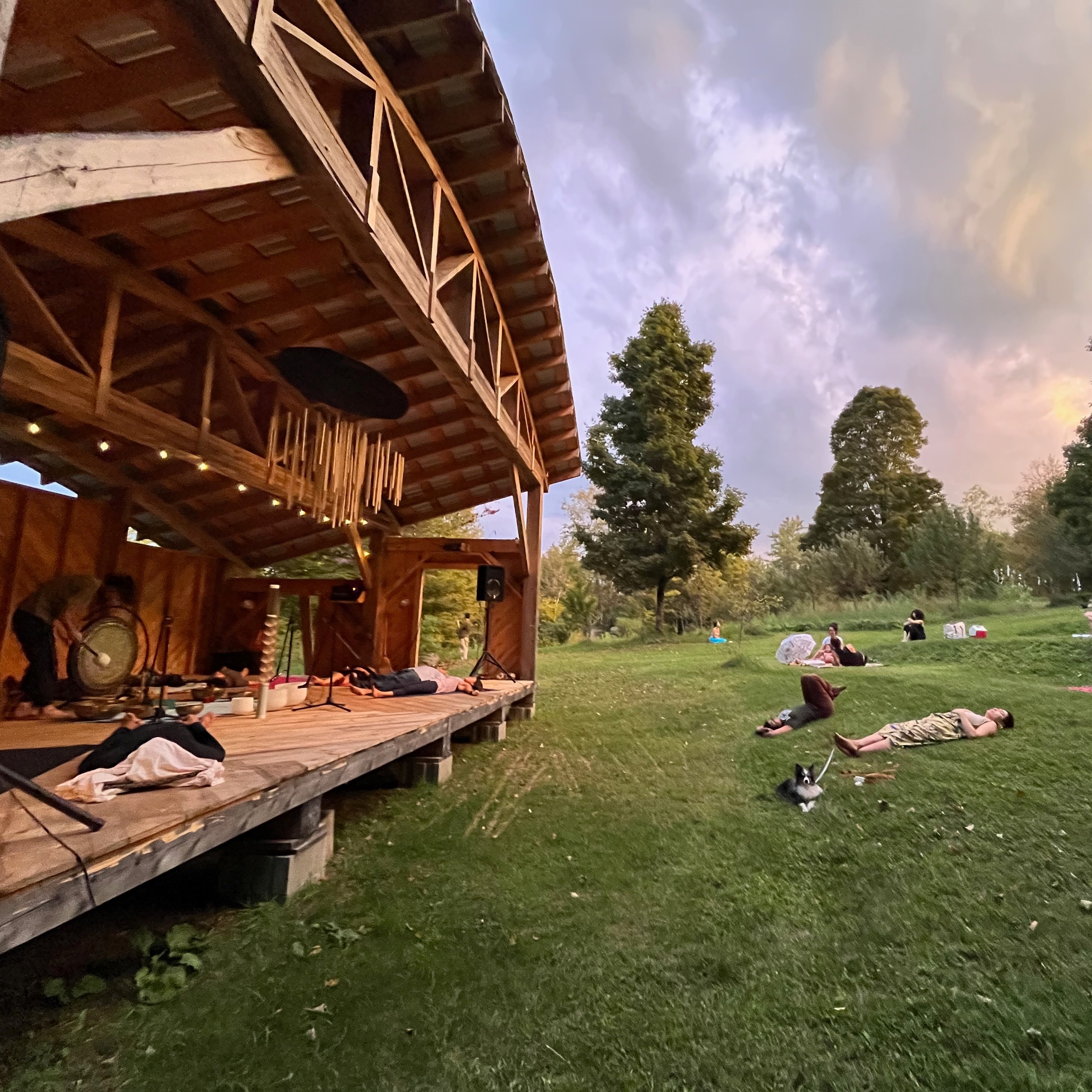 A magnificent wooden stage with a curved roof hosts a Sound Bath: Artist Tony Bednar rings a gong for participants lying on the stage in and in a grassy amphitheater. 
