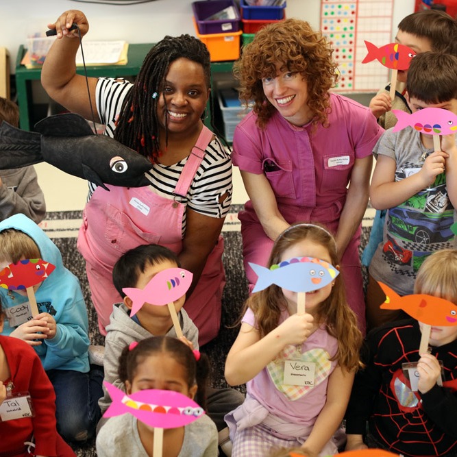 Tanya & Bonnie in a classroom with a fish puppet, surrounded by students