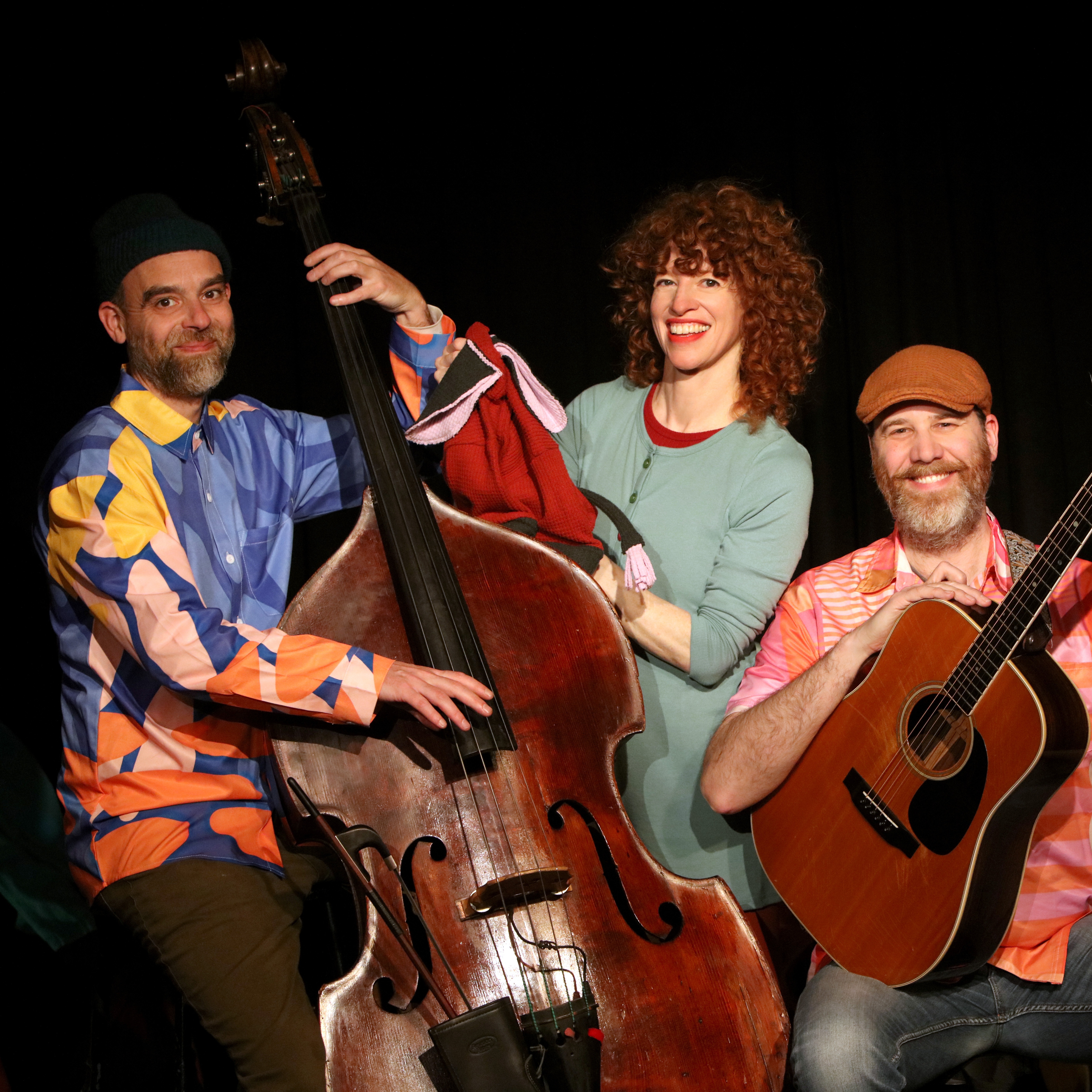 3 white smiling individuals:  a bass player and his instrument; a performer and her puppet; a guitarist with his instrument