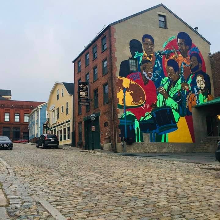 Photo facing Northwest on Centre St featuring Kat Knutsen's "Jazz Wall" Mural outside of the Fiber Optic Center's loading dock in New Bedford that also includes a cobblestone street, a few cars and a few other buildings. 