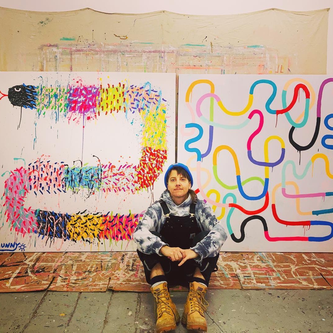 Person sitting on the ground wearing black overalls, boots and a blue hat, in front of two large colorful paintings.