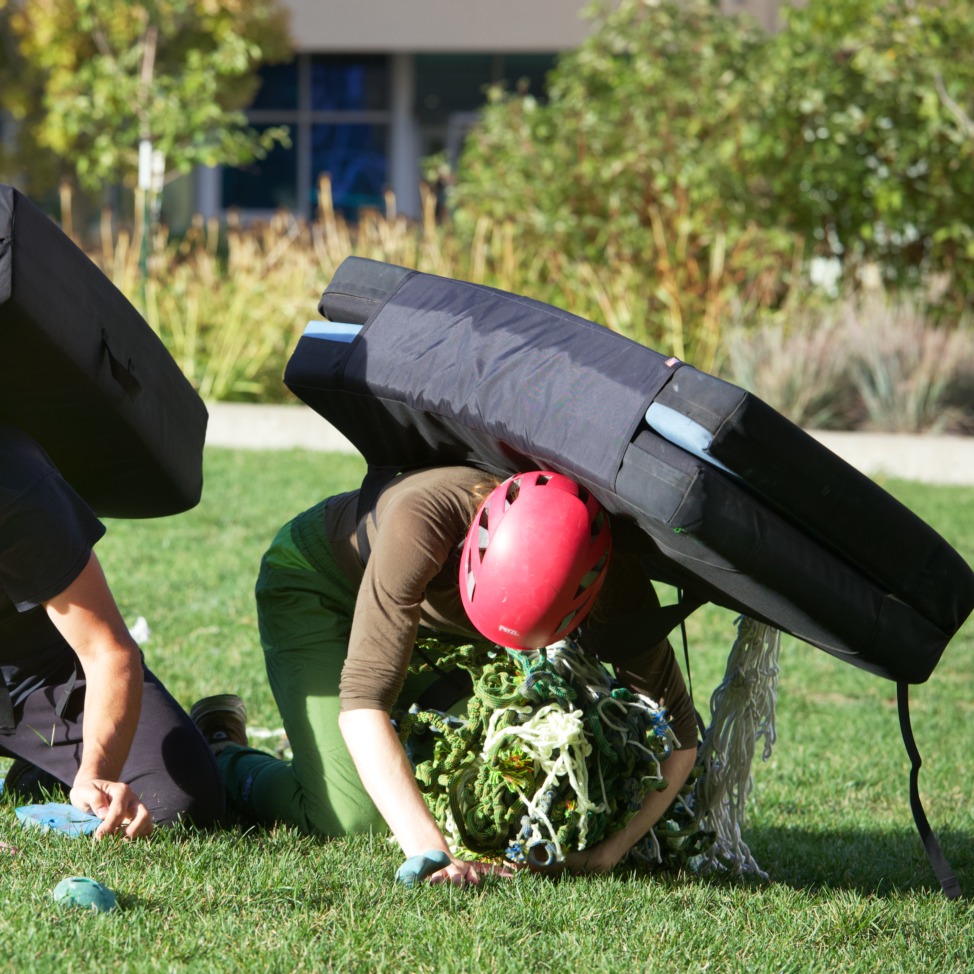 Ellen Oliver is crawling in a green field. She is wearing a climbing crash pad, pink climbing helmet, green pants, and a green shirt. She is hold onto climbing rope.