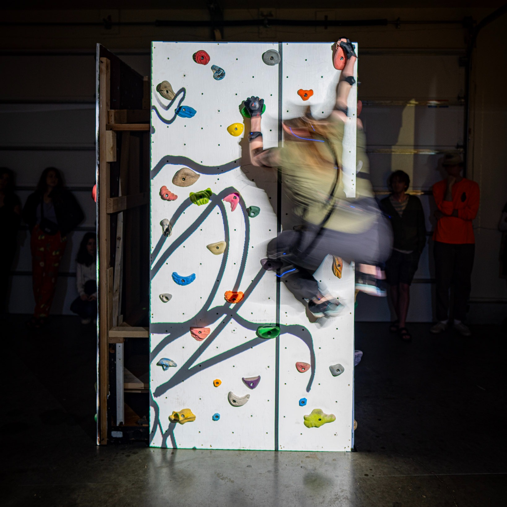 Ellen is wearing a green t-shirt and dark pants. She is climbing on a white climbing wall with curved black lines projected onto her. She is wear motion capture sensors.