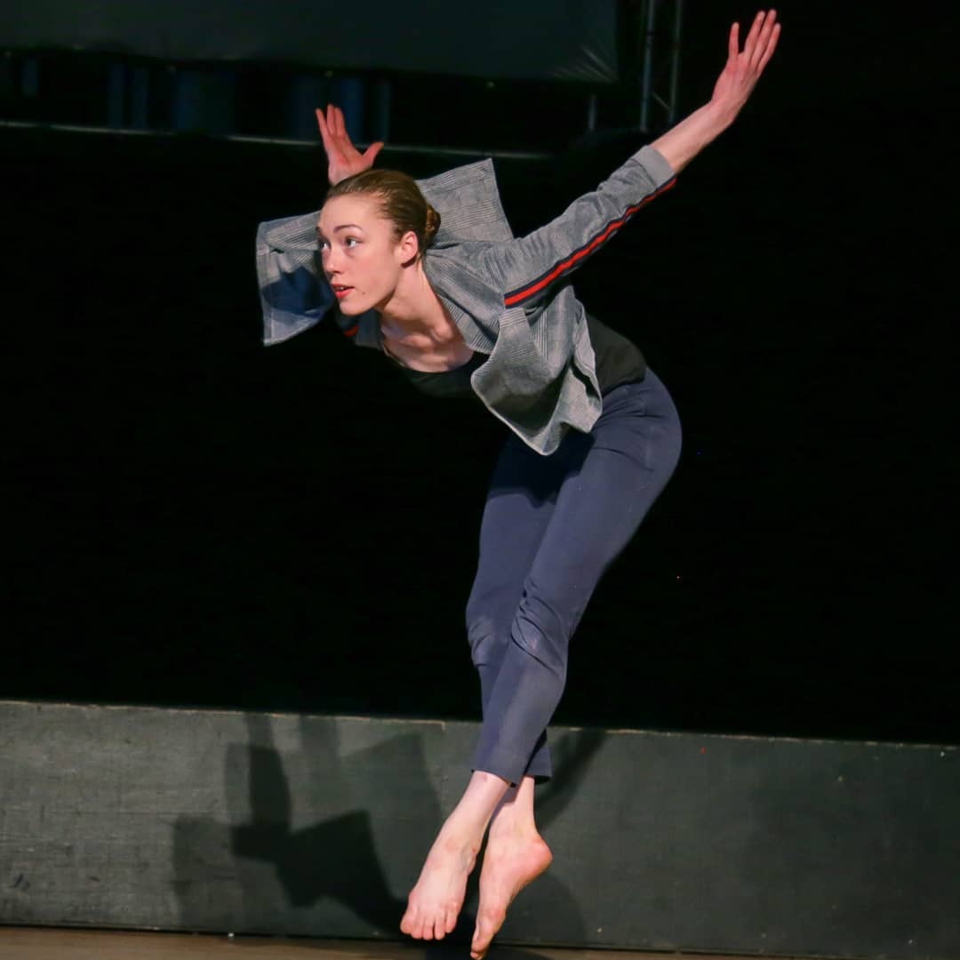 Ellen Oliver is wearing a grey jacket and blue pants. She is in a jump. Her legs are pointed towards the floor as she leans forwards, and her arms are stretch behind her.