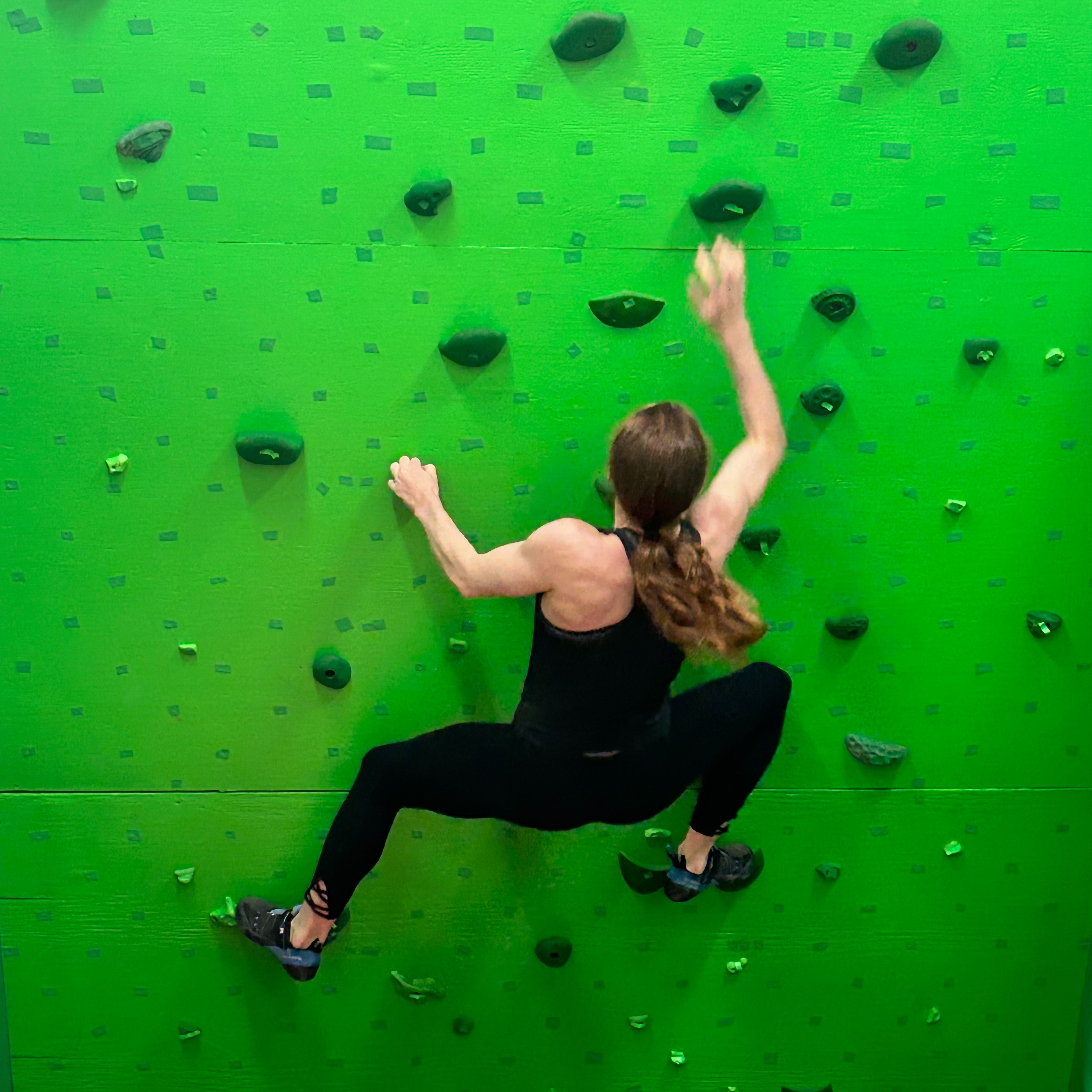 Ellen is dressed in all black and climbing on a green climbing wall. She is reaching her right hand up towards a hold.