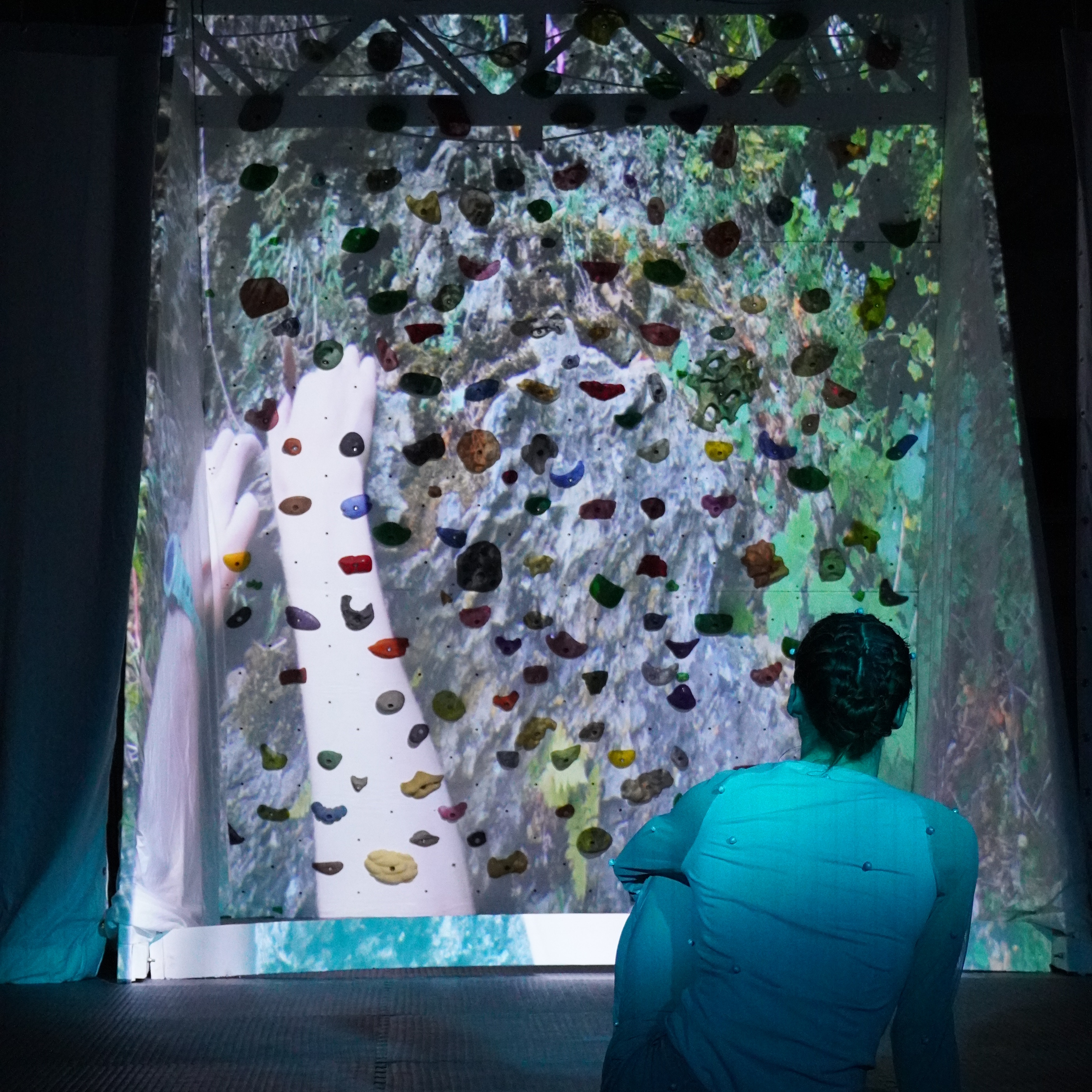Ellen Oliver sitting and looking at a projected image of hands on a climbing wall. She is wearing white, and she is sitting a blue light.