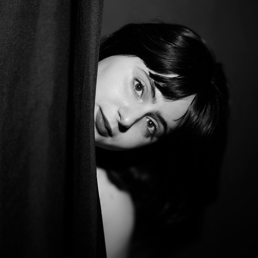 A black and white photo of Mira peaking out from behind a black curtain. Mira has pale skin, dark shoulder length hair, bangs, and a dark lipstick on.