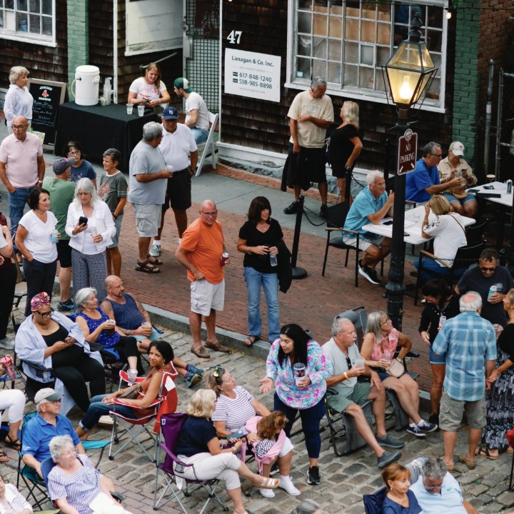 Day time photo from a 2nd story balcony, overlooking lower Union St during Summer Sound Series. The photo includes patrons sitting in camping chairs as well as standing on a cobblestone street and along the sidewalk. 