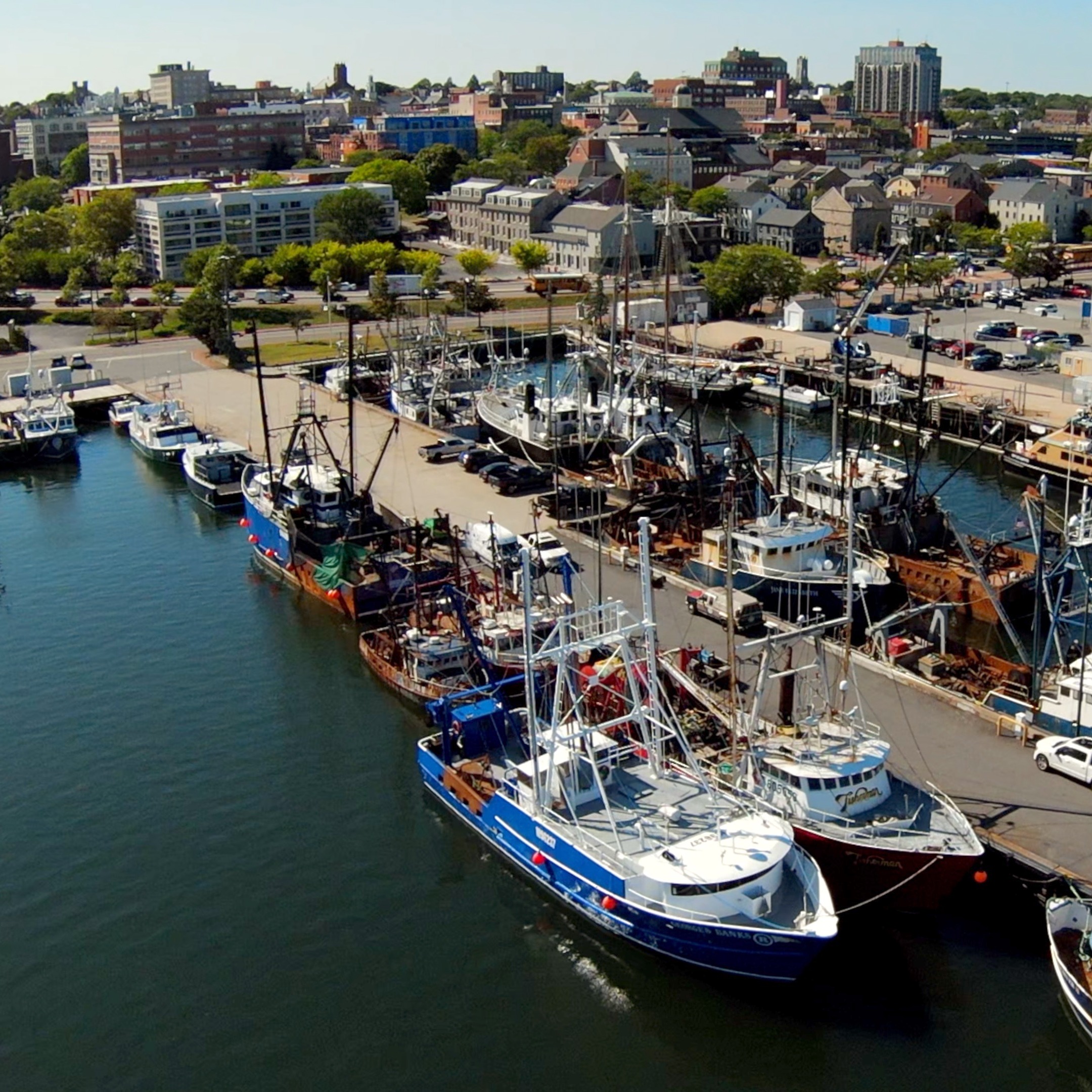 Day time drone photo looking West from the Port of New Bedford. Photo includes various commercial fishing vessels as well as a docking area for the ships. There are buildings in the background