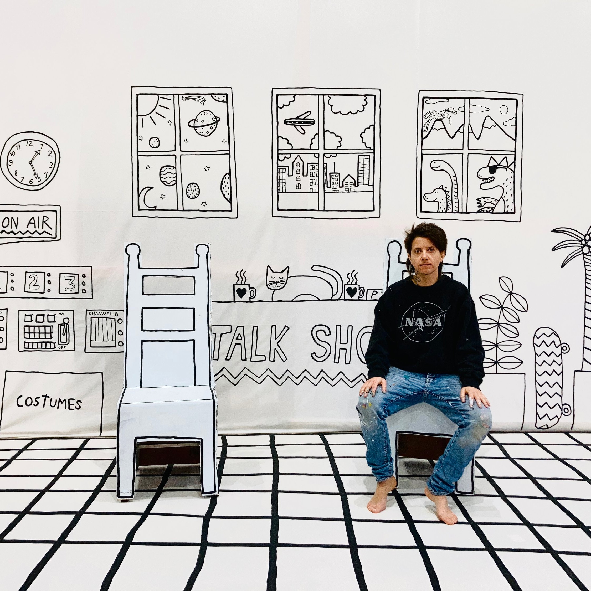 Photo of a person sitting in a cardboard chair in a black and white talk show set