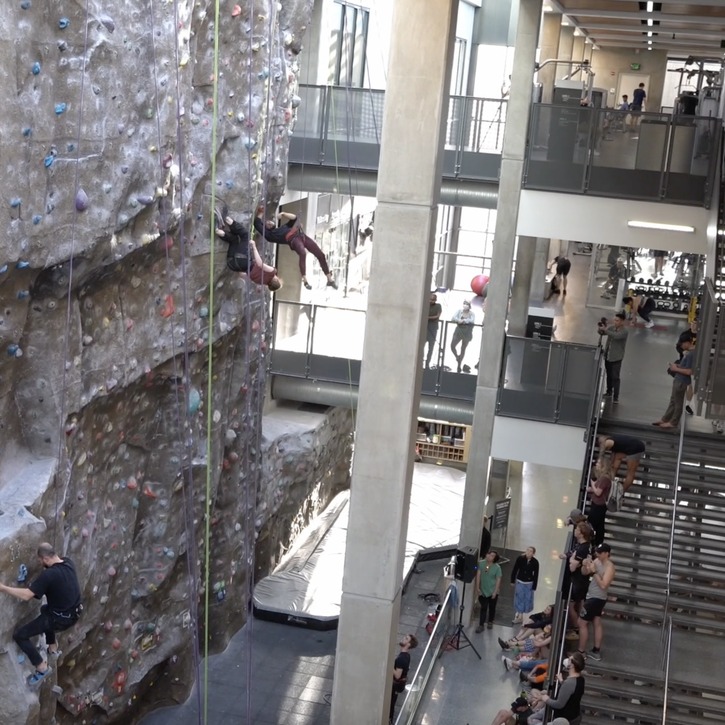 Ellen and Raud are wearing black and red. They are dancing on a climbing wall, in front of a staircase with audience. Ellen and Raud lean backwards away from the wall as they are suspended with ropes and harness.