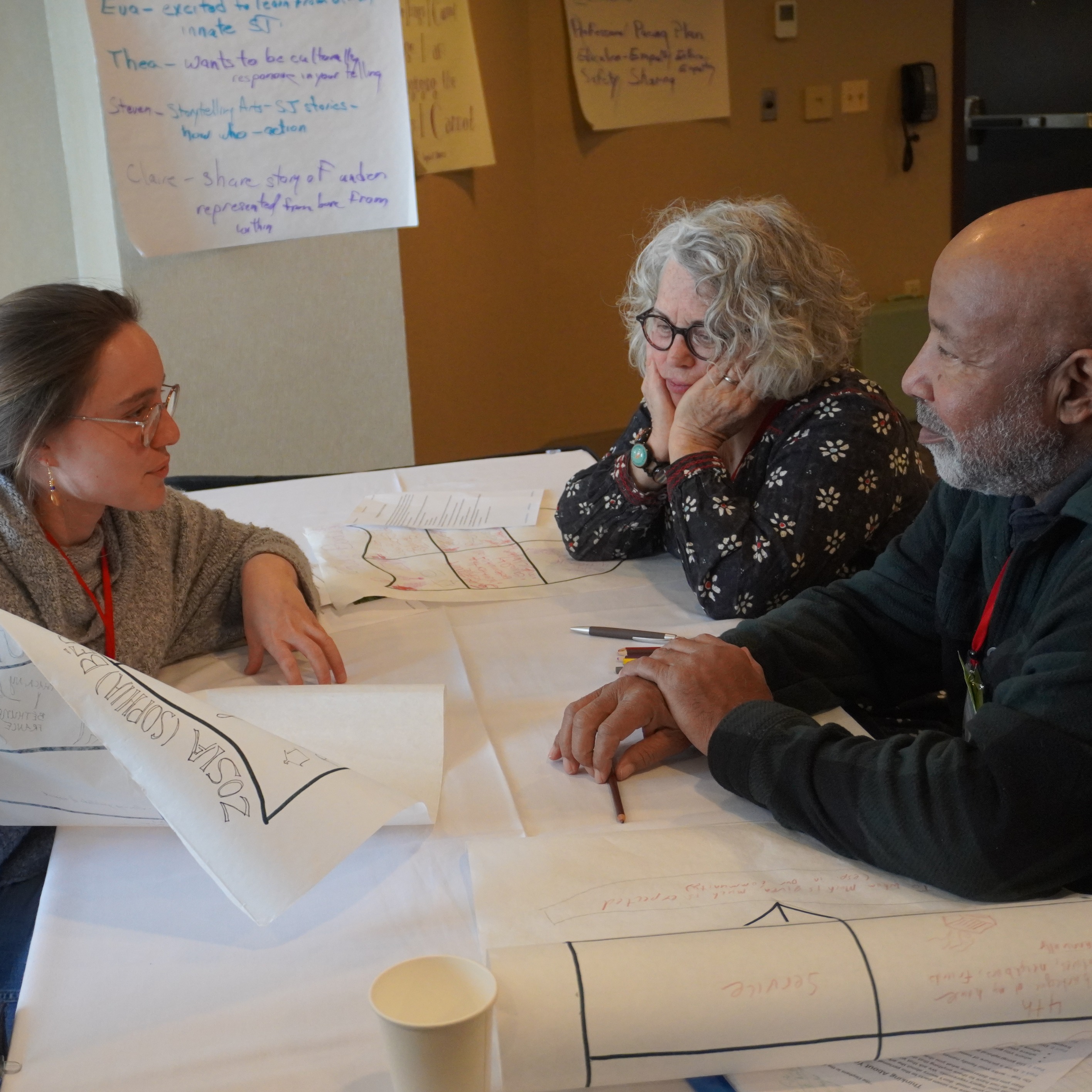 Three storytellers engage in active work during a workshop. A young woman with glasses holds a large piece of paper. Pens sit on the table between two older individuals. 