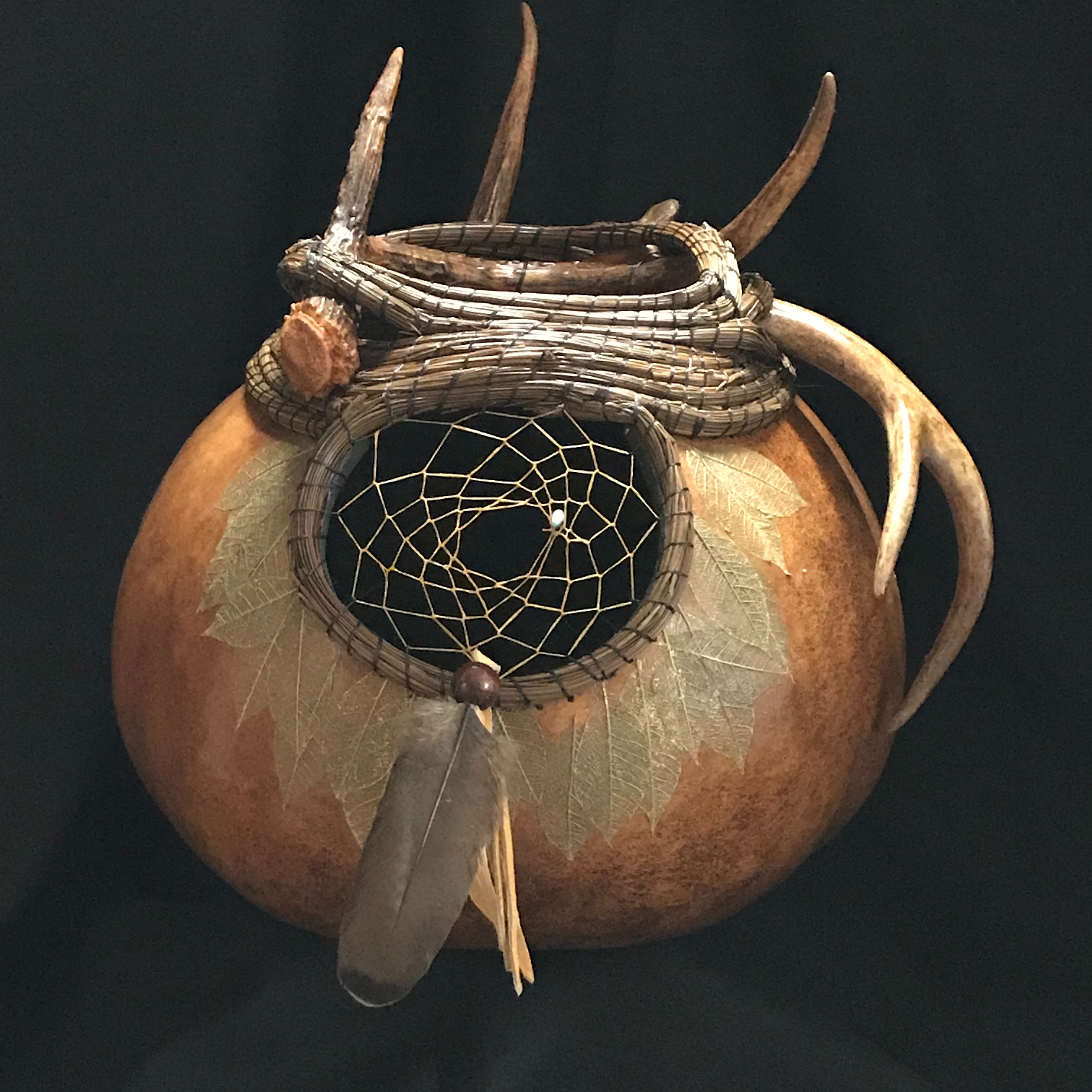 Larger gourd featuring pine needle coiling and white taile deer antler.  The dream catcher is surrounded by skeletonized leaves and accented by a duck feather and deer hide fringe.
