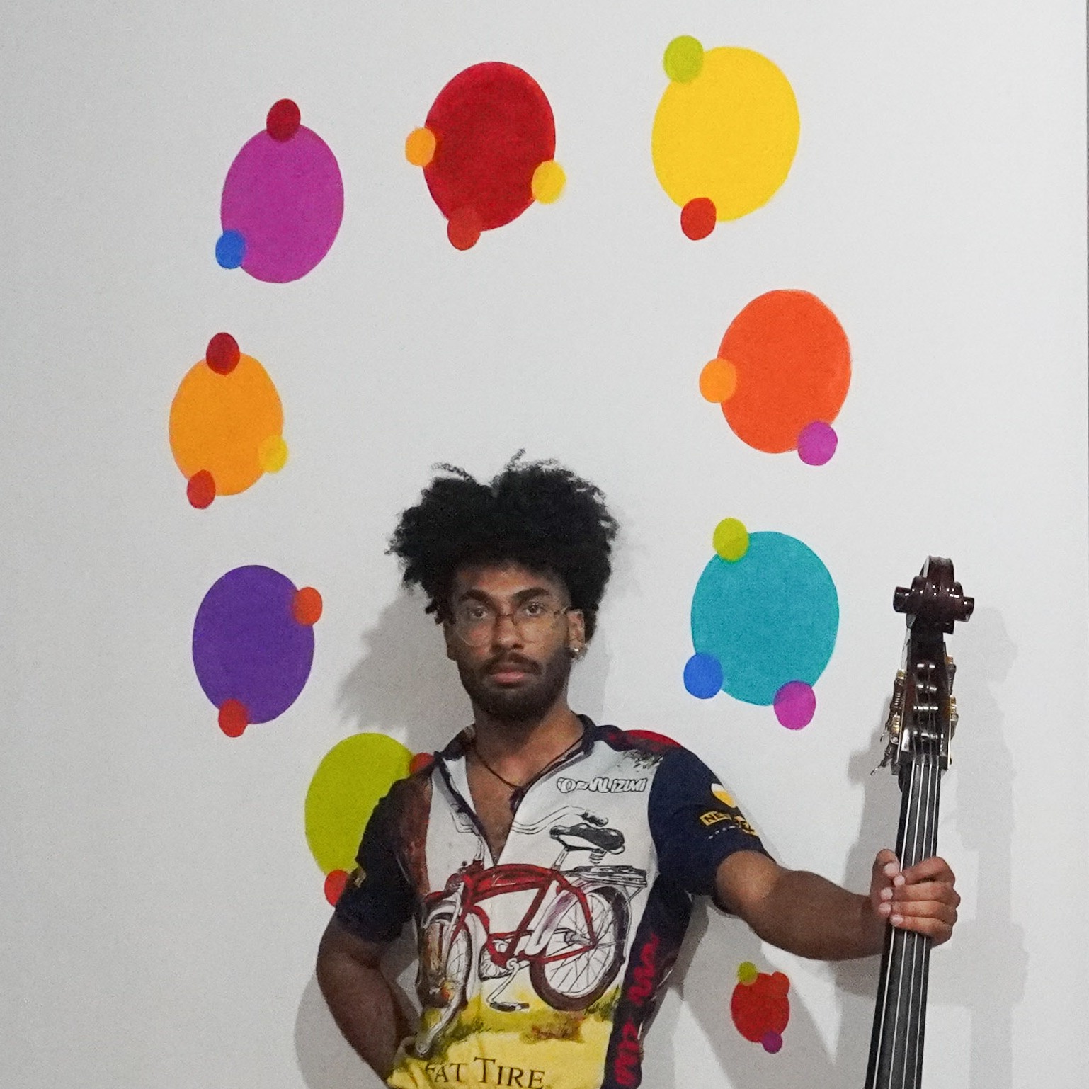 Indë holds their double bass and stands before a mural depicting a chord progression. On the wall to the right is their painting "Marsha P. Johnson as The Three Ages of Woman, after Gustav Klimt." 