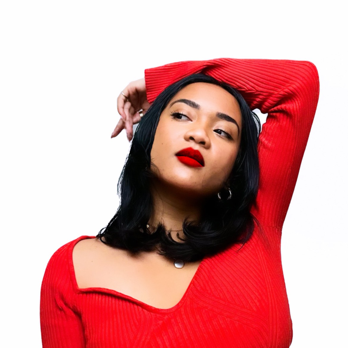 A portrait of Aurora in a red long-sleeve top and bold red lipstick, with black, straight, shoulder-length hair. Her arm is raised above her head, and she gazes to the right against a white background