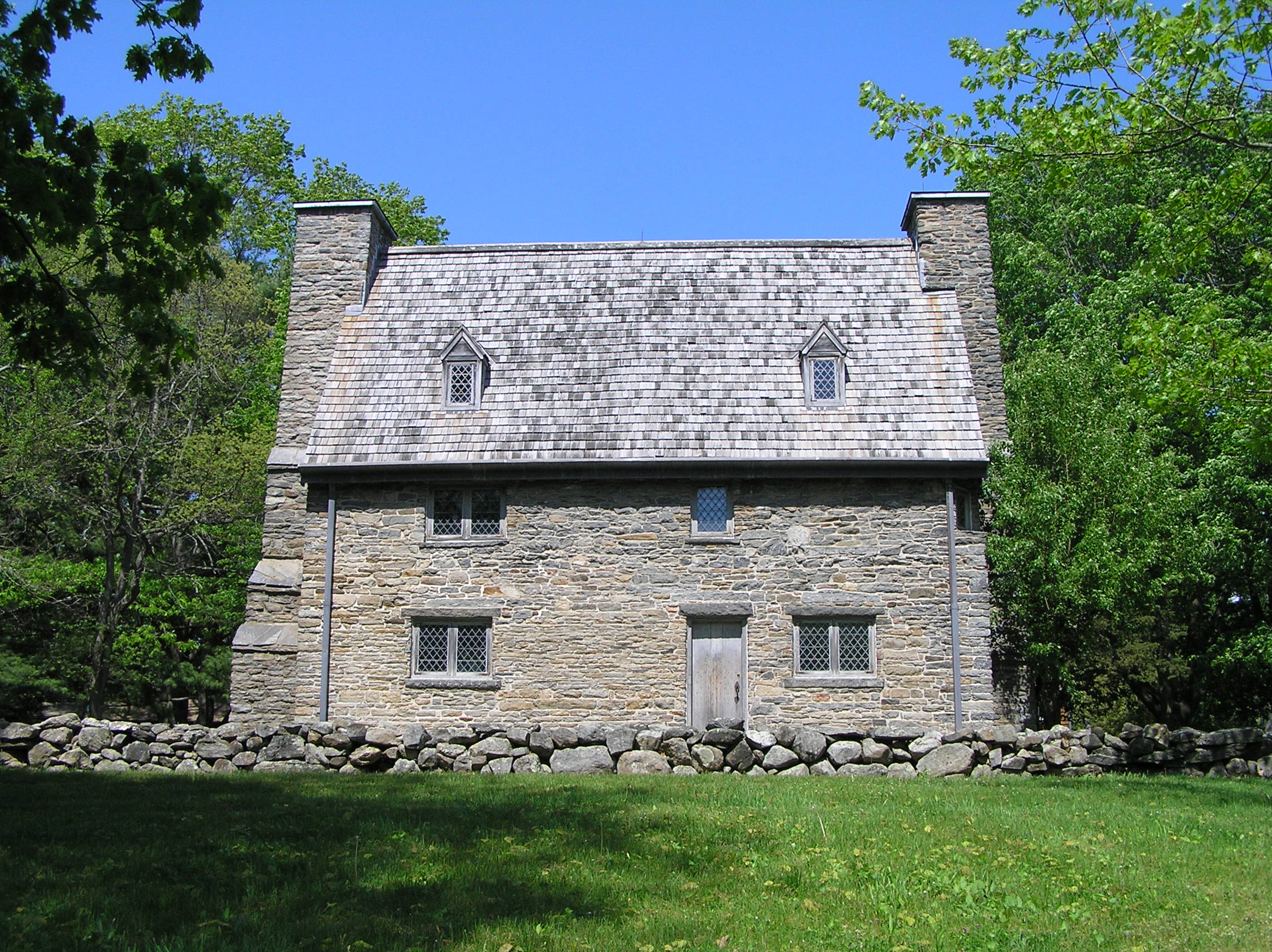 2-story gray granite stone house with a steep wood-shingled roof and a large chimney stack at left and right