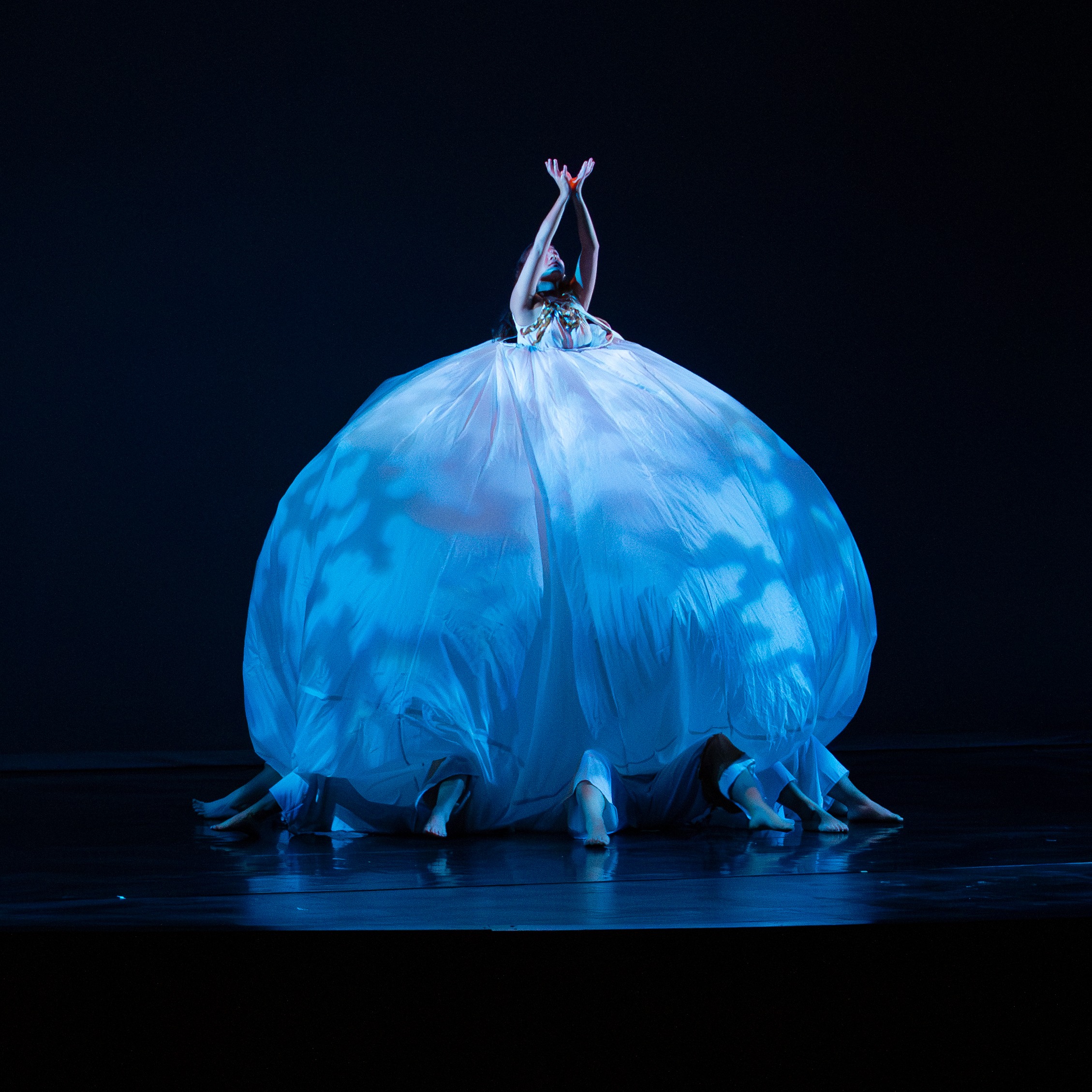 woman in large white skirt with multiple dancers bent legs sticking out form underneath the skirt