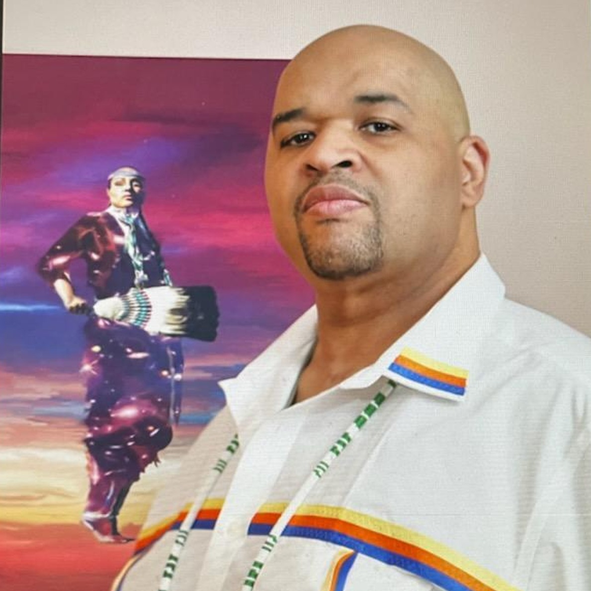Headshot of Justin Beatty wearing a white Native American ribbon shirt and beaded necklace standing in front of a piece of his artwork titled "Universal Healthcare".