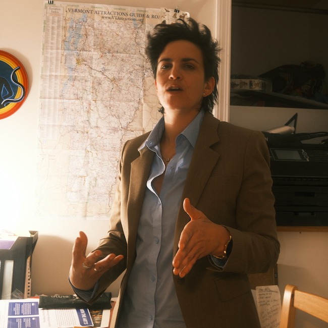 A photo of Rachel Lindsay in her studio, speaking about her work to a photographer. She is wearing a beige blazer and blue collared shirt, the top two buttons undone. In the background, the bright light of a lamp blows out the lower part of  a VT road map