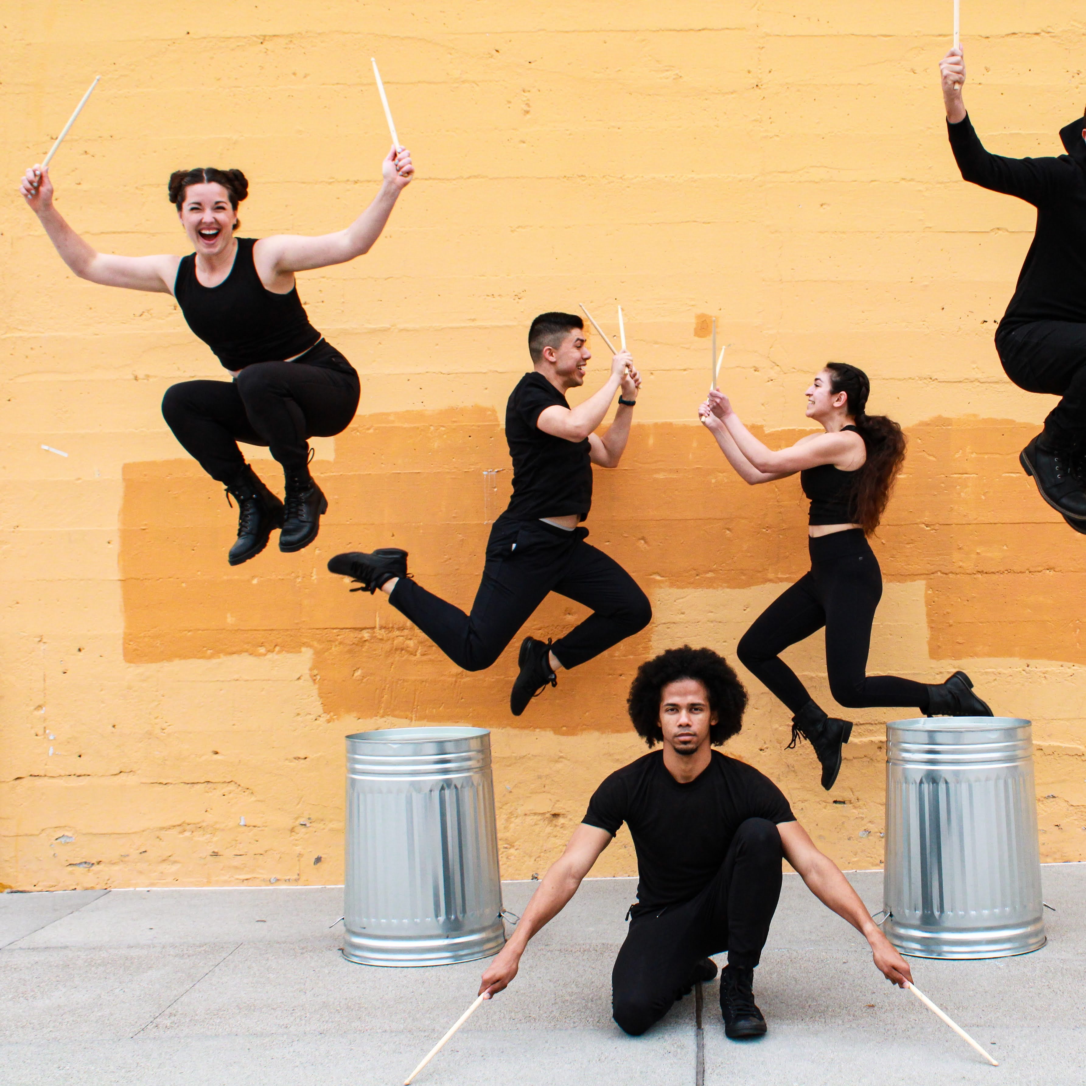 A group of performers dressed in all black, jumping in the air holding drumsticks, one dancer kneeling on the ground with drumsticks hitting the floor.