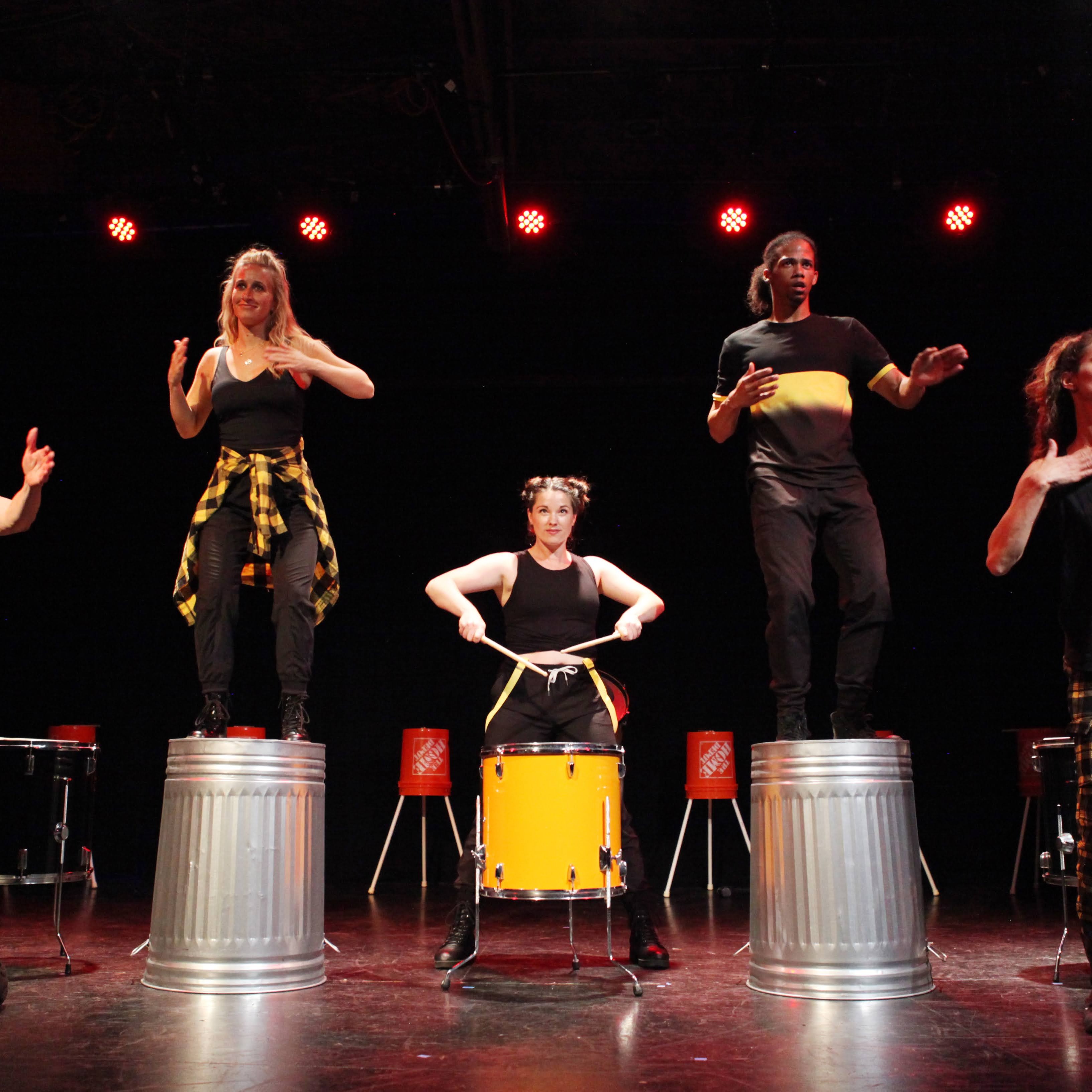 Three performers dressed in black and yellow, two standing on top of aluminum barrels and one drumming behind an orange drum