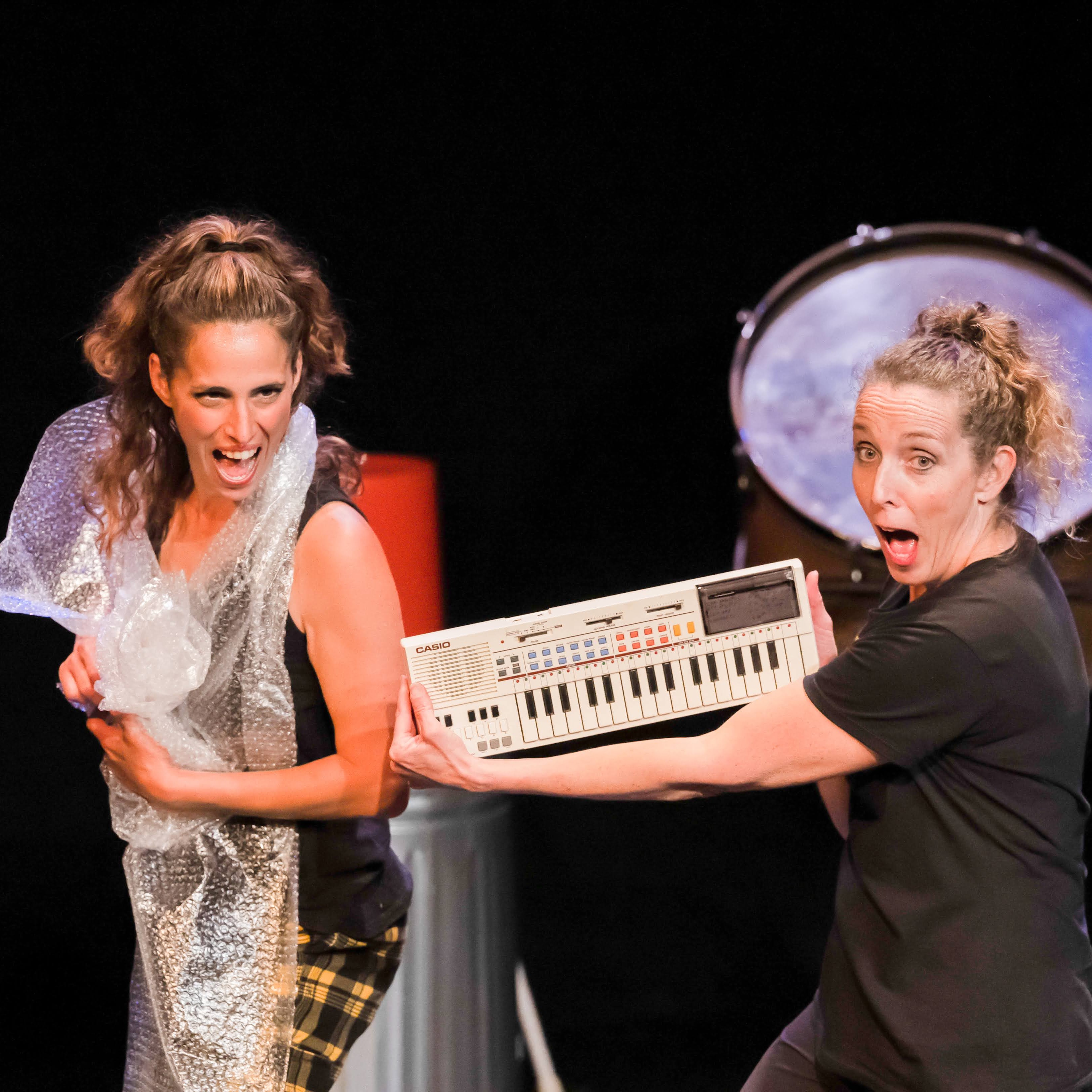 Two performers, one holding a vintage Casio keyboard, the other wrapped in bubble wrap, both with surprised expressions.