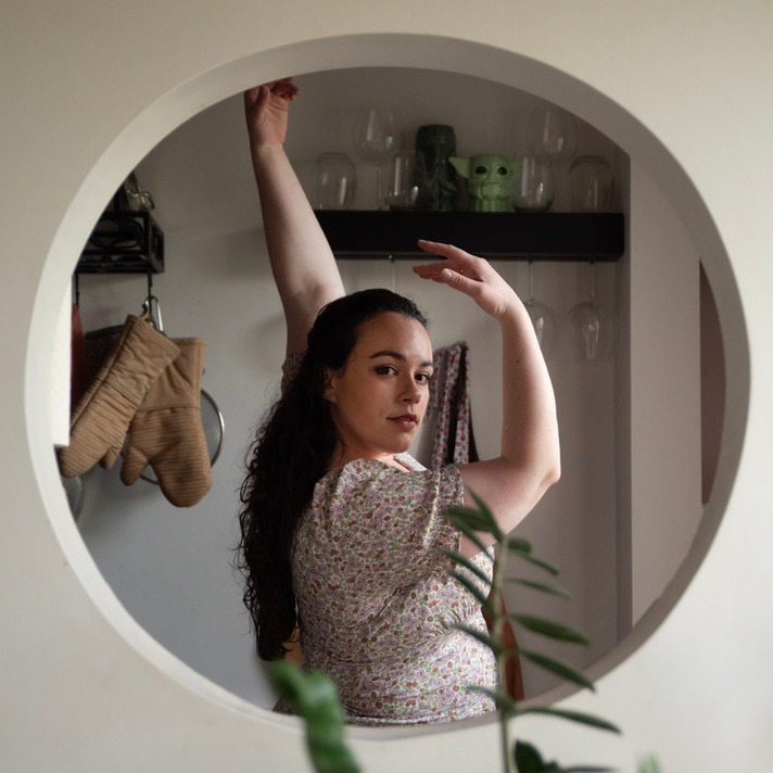 A white woman in a floral dress with long curly dark hair has her hand up near her face while looking through a circular 