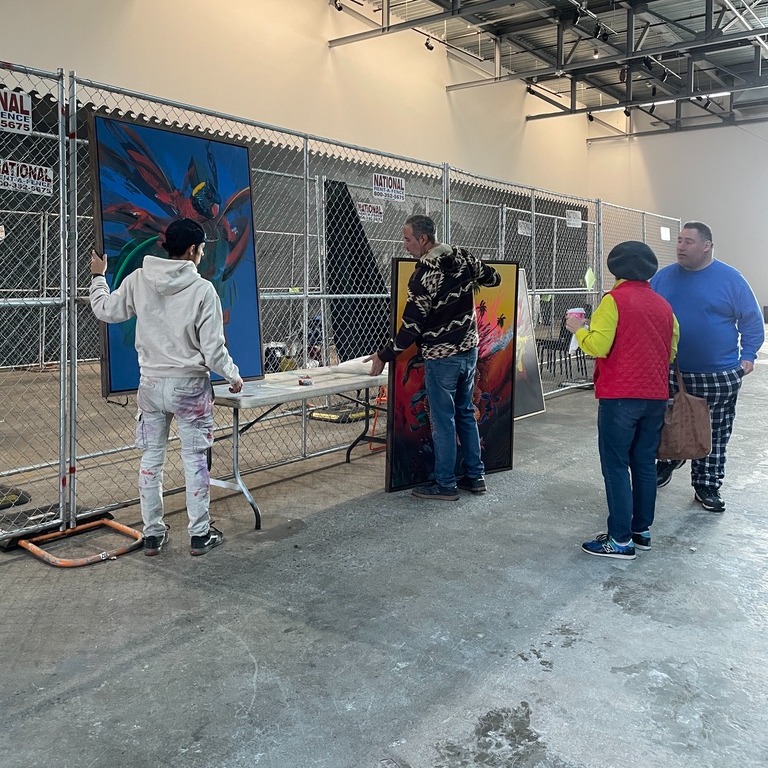 Four people gathered near chain link fence, inside large industrial space. Two are holding large, colorful paintings, ready to hang on fences. 