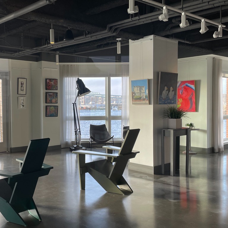 View of Boston East Gallery with artwork hanging on walls. Chairs and oversized floor lamp that looks like a desk lamp fill the floor space. Window in background overlooks part of Boston Harbor, facing Chelsea, MA.