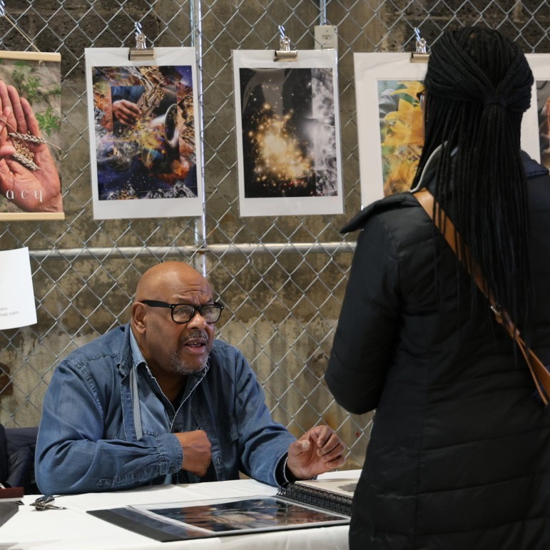 Man with black eyeglasses and blue shirt sitting at table with a book of photos, and photos hanging on chain link fence behind him. He is speaking with a woman whose back we see. She is wearing a black coat.
