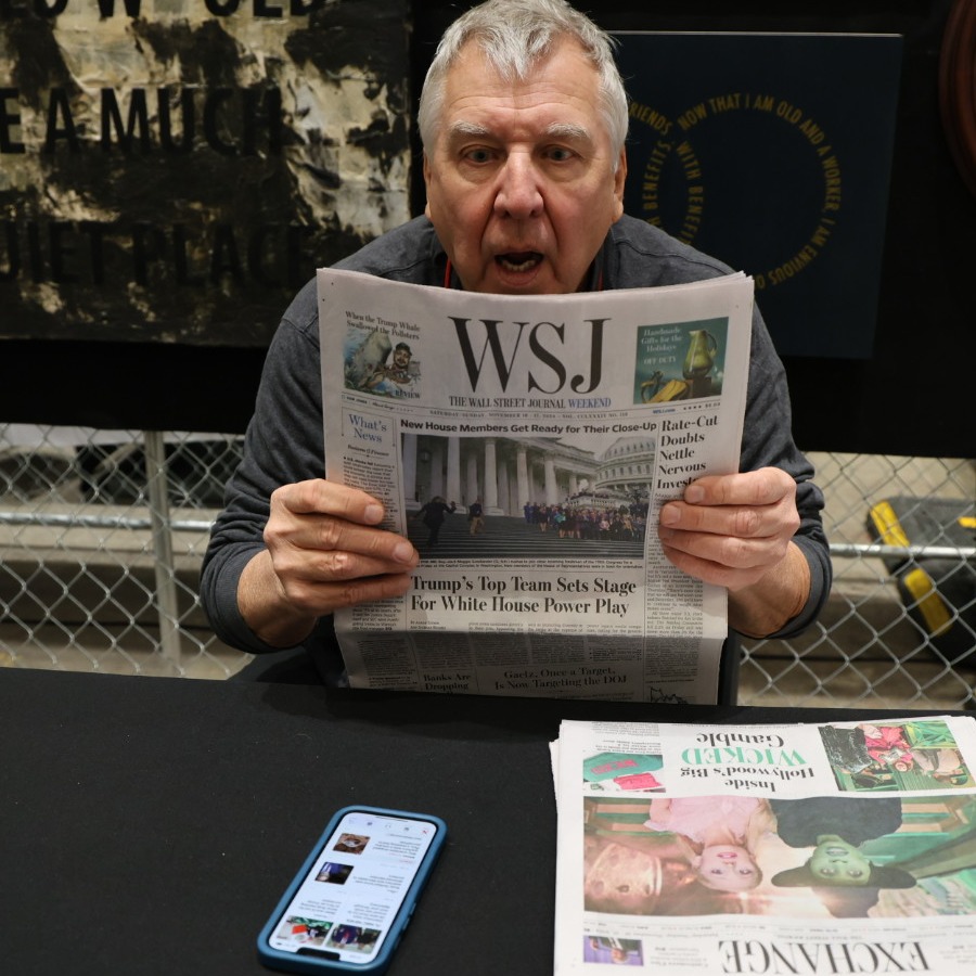 Man in gray shirt, sitting at a table, holding a copy of the Wall Street Journal. His mouth is agape and he looks surprised. Dark, text-based art hangs behind him on a chain link fence. A cellphone showing text and images is open in front of him.