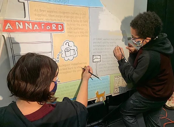 Two middle school students wearing masks write with pencils on a wall mural